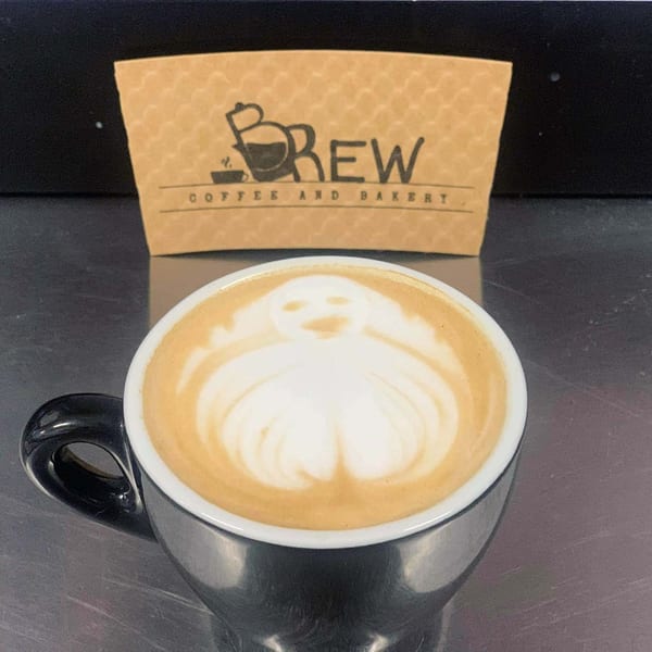 A black coffee cup with latte art depicting a ghost, with a "Brew Coffee and Bakery" sleeve behind it.