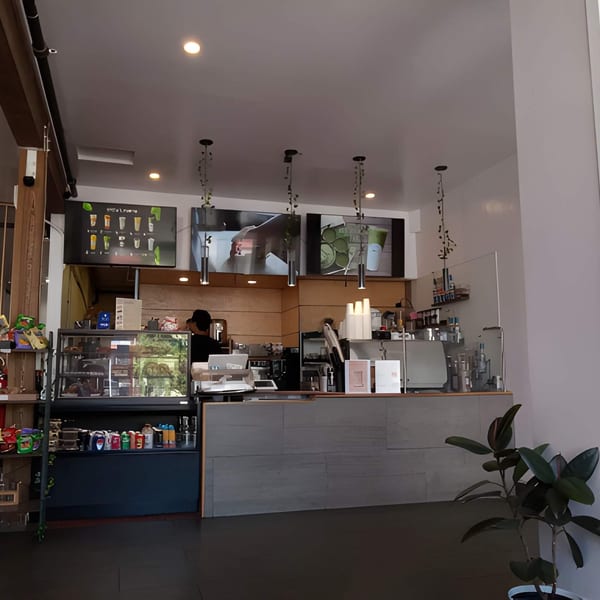 Interior view of a cafe with a counter, display cases, menu screens, and a plant.