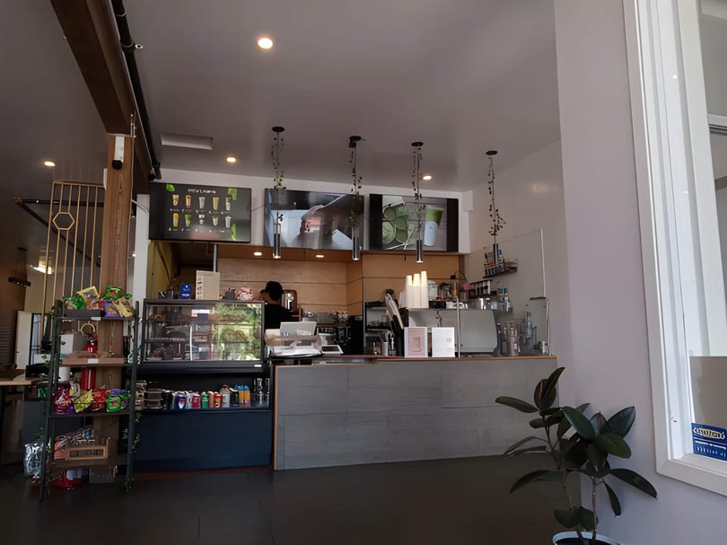 Interior view of a cafe with a counter, display cases, menu screens, and a plant.