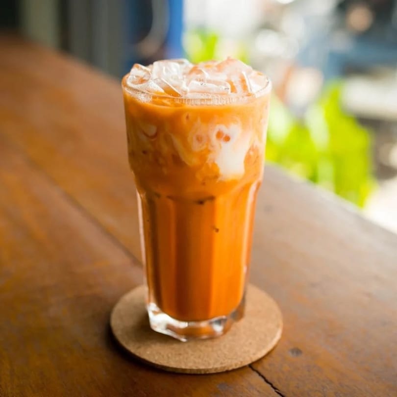 Iced Thai tea with milk swirls and ice in a tall glass on a wooden table.