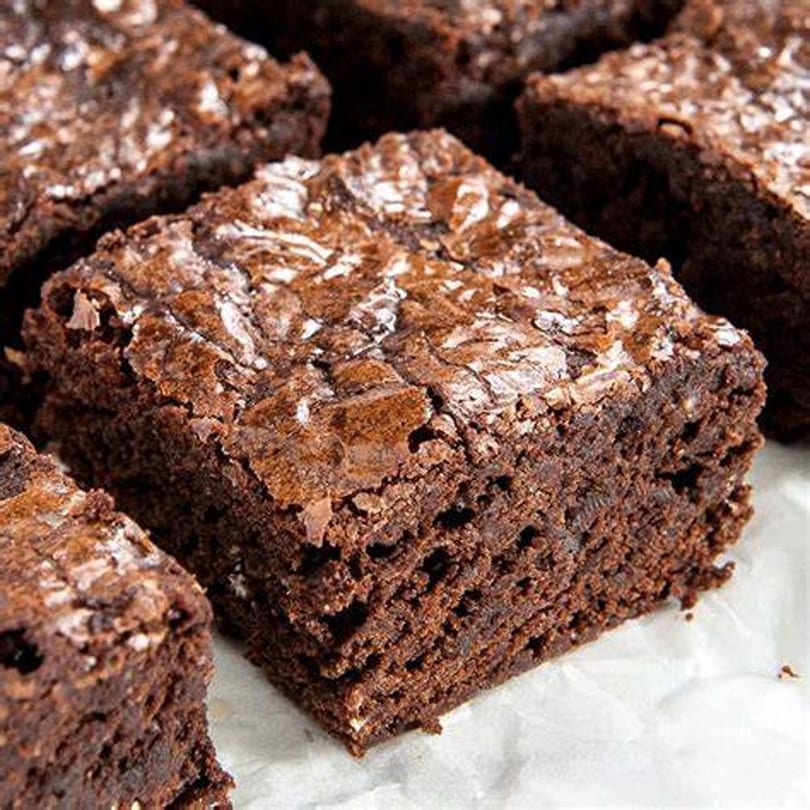Close-up of a rich, fudgy chocolate brownie with a crinkled top, on white parchment paper.