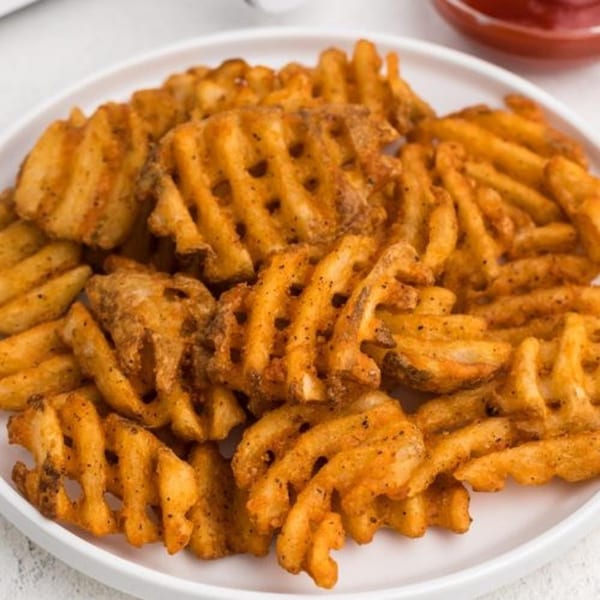 A white plate piled high with golden-brown, seasoned waffle fries, with a small dish of ketchup in the background.