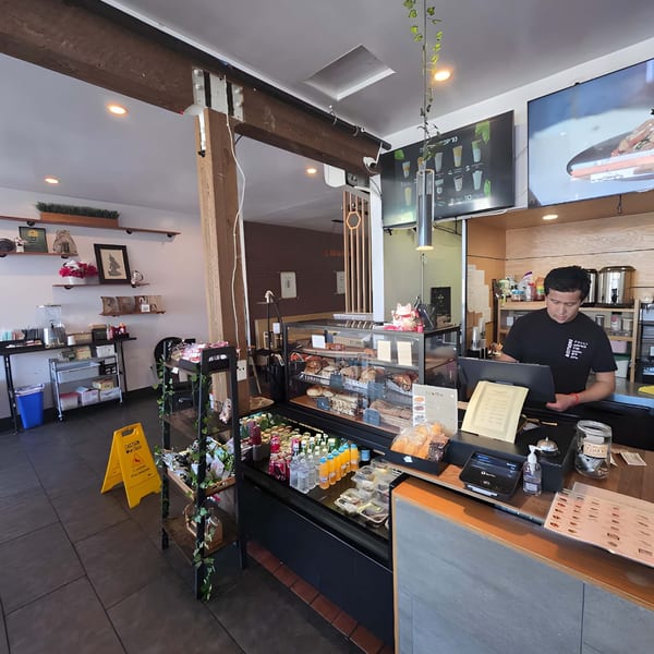 A man stands behind a counter with pastries, drinks, and a cash register in a cafe.