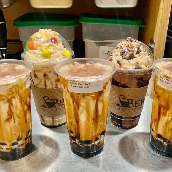 Five boba drinks, three brown sugar boba, one with candy, and one chocolate, on a metal counter.