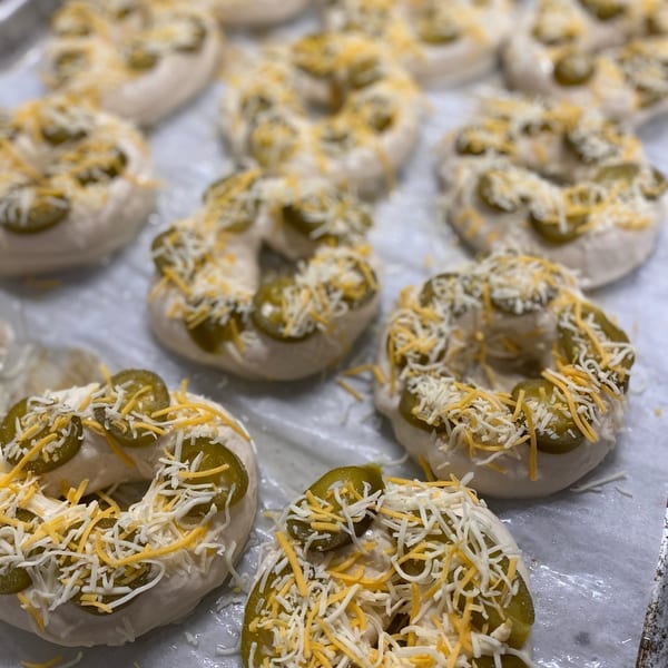 Unbaked bagels topped with sliced jalapeños and shredded cheese on a baking sheet.