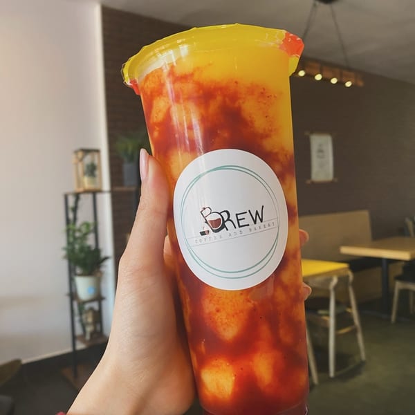 Hand holding a Brew Coffee and Bakery mango drink with red chili swirls inside a cafe.