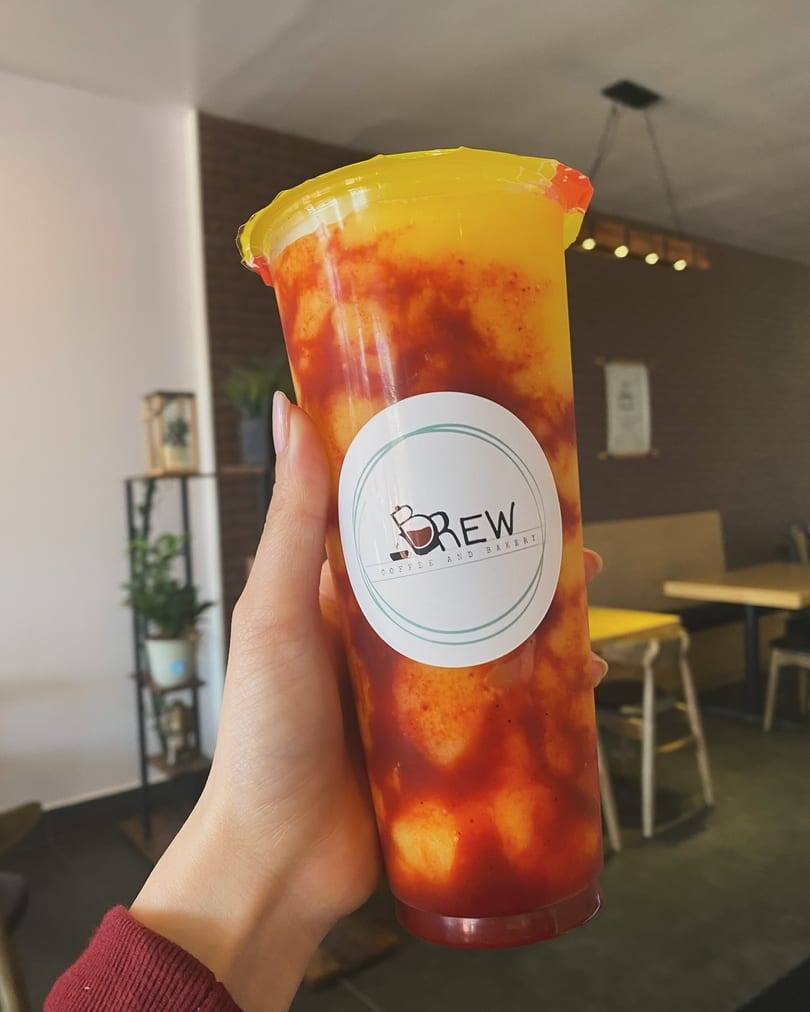 Hand holding a Brew Coffee and Bakery mango drink with red chili swirls inside a cafe.