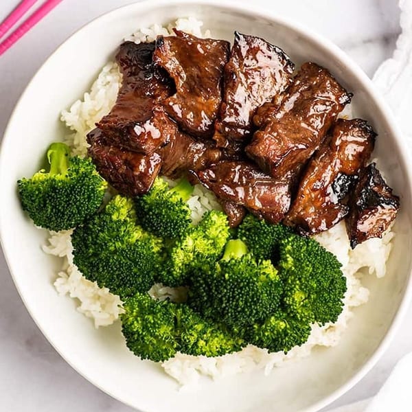 Teriyaki beef and broccoli served over white rice in a white bowl with pink chopsticks.