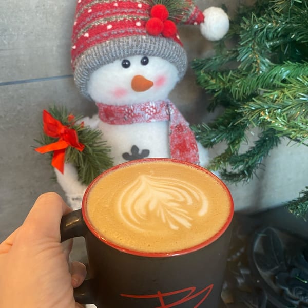 Hand holding a black mug with latte art, next to a snowman decoration and a Christmas tree.